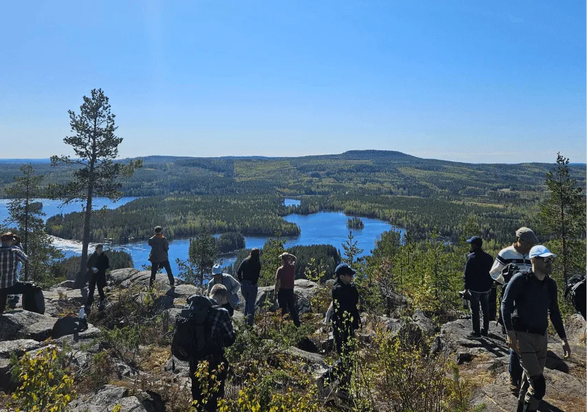 Hikers gather at the top of a sun-drenched hill with lakes and woodland in the background below