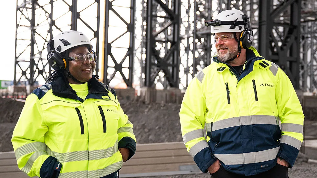 Two colleagues in safety equipment, including hard hats and reflective clothing, stood together on a job site