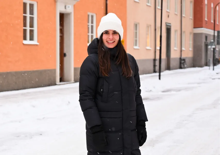 A woman in a long black puffer jacket stands in the snow on a Swedish street