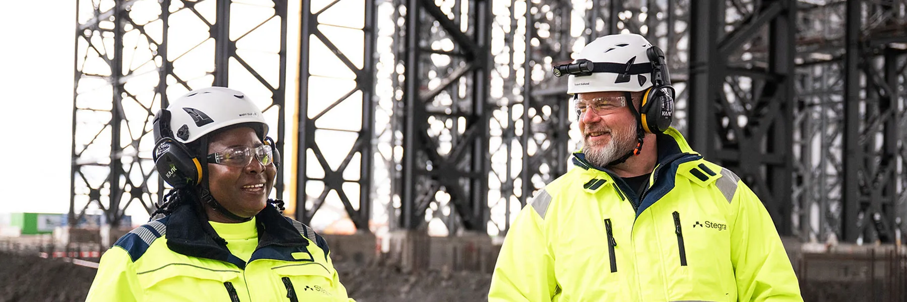 Two colleagues in safety equipment, including hard hats and reflective clothing, stood together on a job site