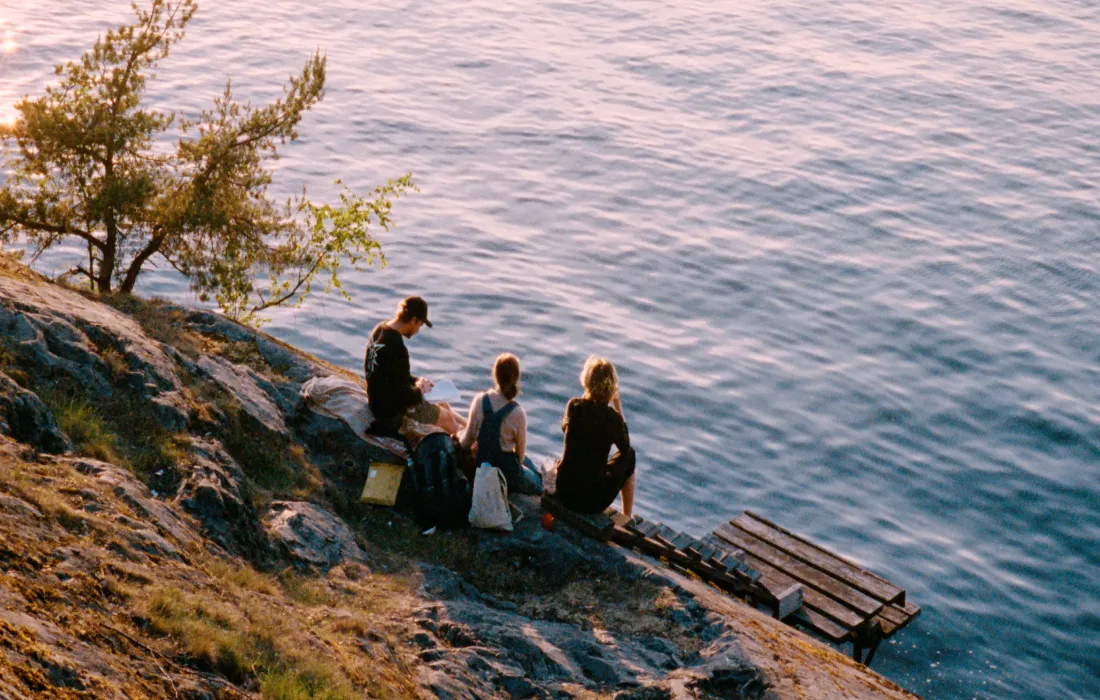 Three people sit on a rock by lake water