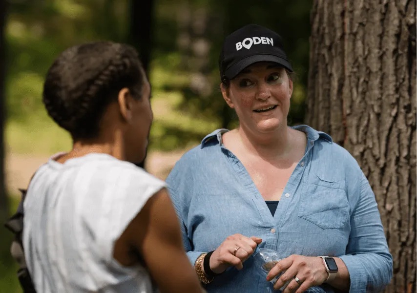 A woman wearing a Boden cap converses with another woman in woodland